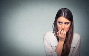 Young hesitant nervous woman biting her fingernails craving something or anxious, isolated on gray wall background with copy space. Negative human emotions facial expression feeling