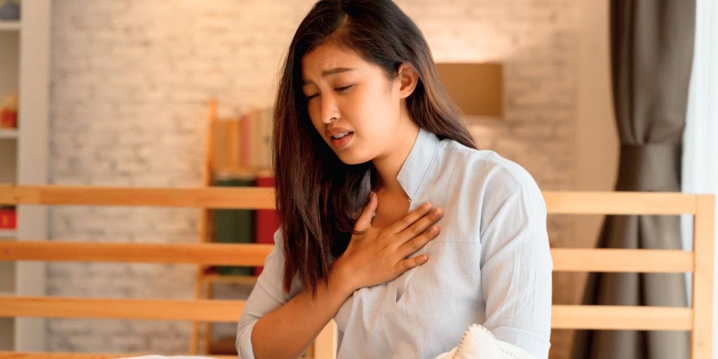 A woman sitting indoors holds her chest with a concerned expression, appearing to experience discomfort or chest pain.