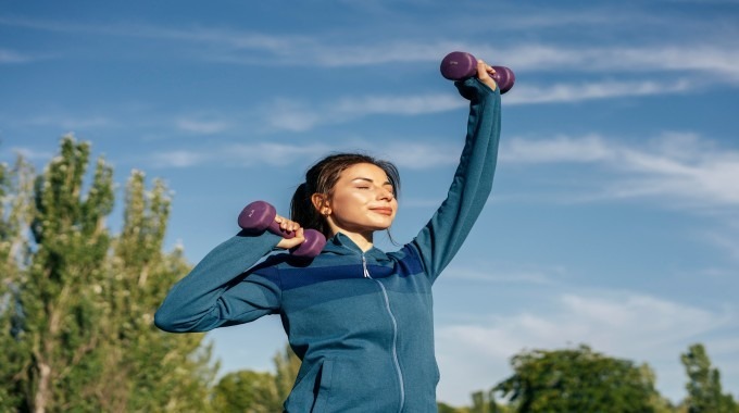 Woman exercising outdoors while lifting dumbbells, promoting fitness and wellness.